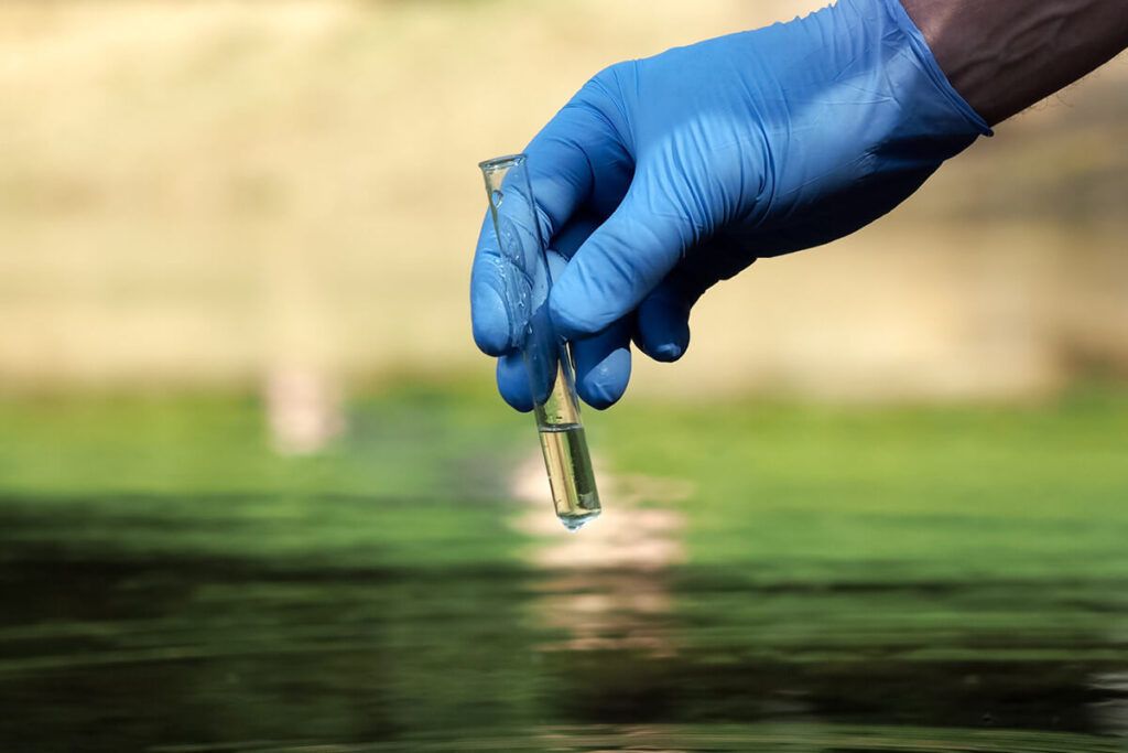 a blue latex glove covered hand collecting a water sample from an outdoor supply for testing