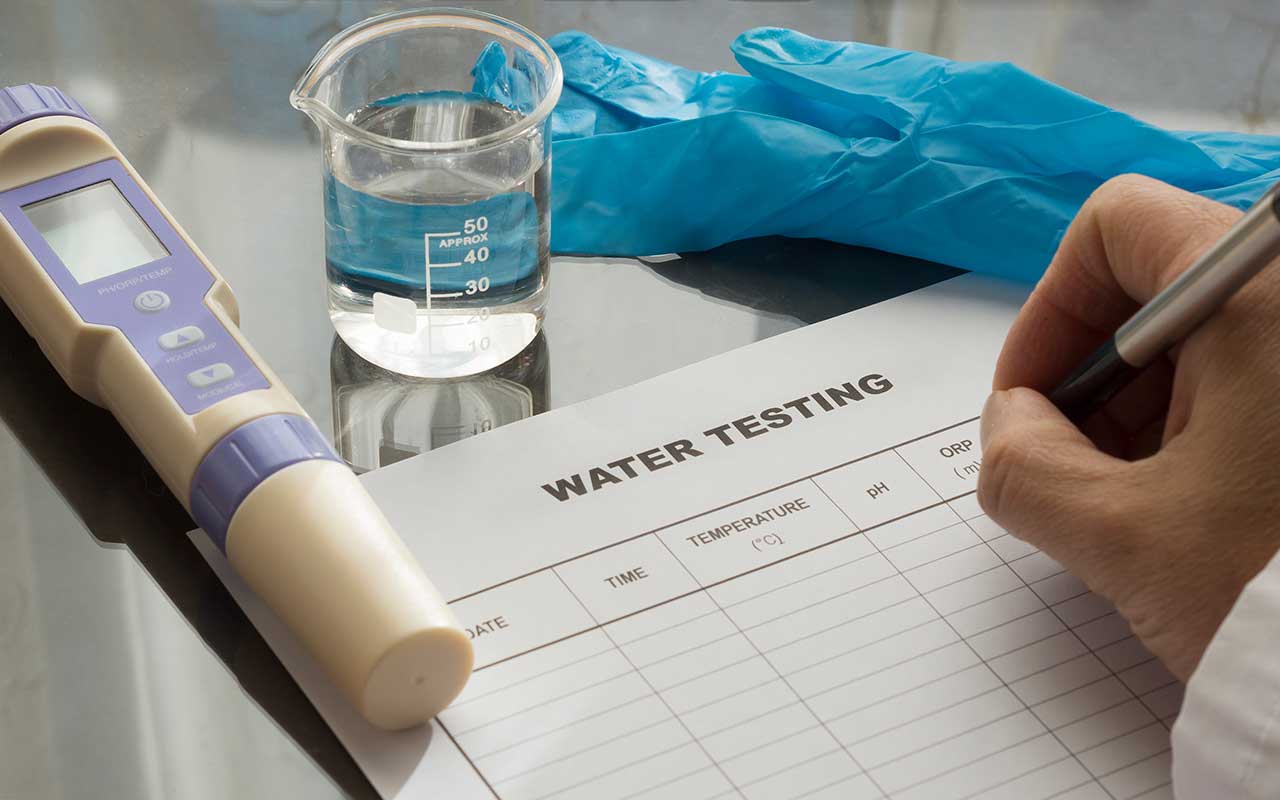someone filling a water testing checklist. A sample of water, latex glove and electronic testing device also on the table.